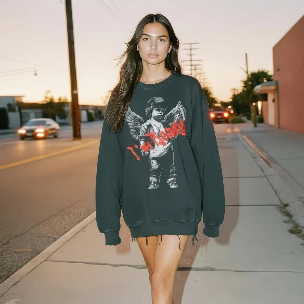 Unisex oversized long-sleeve t-shirt in a black snow wash, crafted from 100% premium cotton. This streetwear tee features a graphic of a cherub or angel with wings, wearing a baseball cap and holding a spray paint can. The design is overlaid with red graffiti-style text. A distressed raw edge hem and frayed details give it a lived-in, vintage feel perfect for a grunge or urban aesthetic
