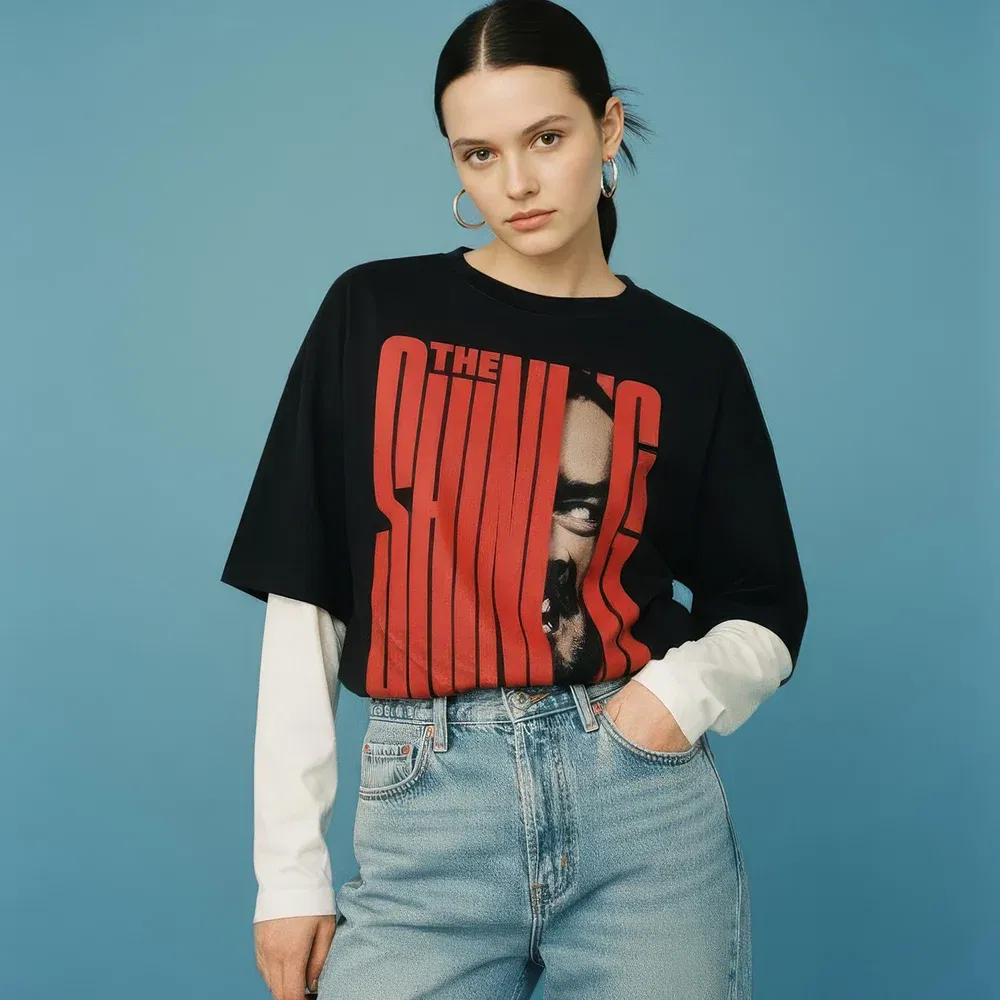 Unisex faux-layered long-sleeve t-shirt made from 100% breathable cotton. This cropped tee features a black short-sleeve layer over white long sleeves, creating a modern streetwear silhouette. The front has a large, iconic graphic from 'The Shining,' with the movie title in bold red text and Jack Torrance's face peering through. A perfect piece for fans of horror movies, 90s style, and urban fashion.
